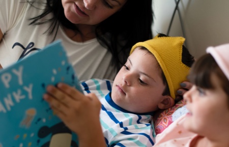 Young child reads picture book in bed with parent and sibling either side of him. His facial features indicate Down Syndrome